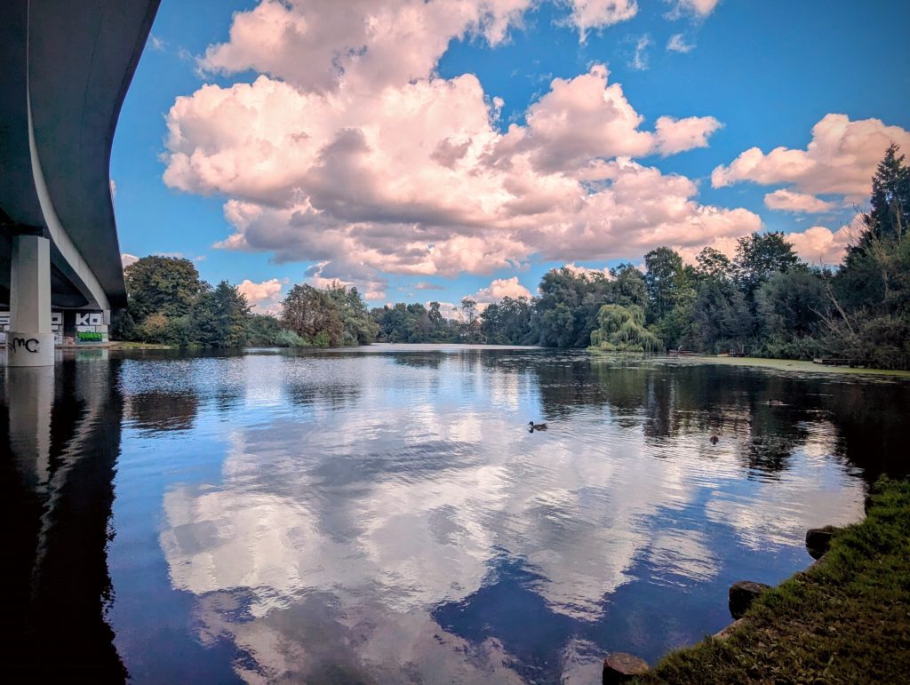 Wakenitz mit gespiegelten Wolken, Bäume an den Ufern, ein Teil der Wallbrechtbrücke, im Lübecker Stadteil St. Jürgen
